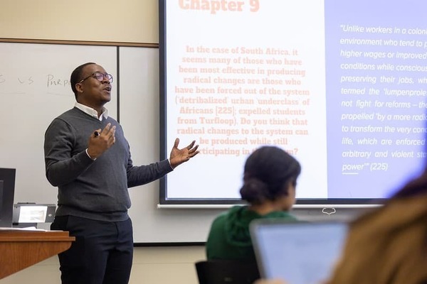 Professor Forjwuor standing at the front of the class next to a screen displaying a power point slide with some text
